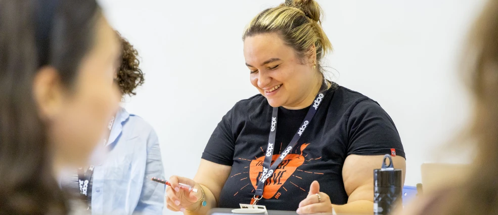 Woman with blonde hair in a bun, wearing a black t-shirt and ECPR lanyard, smiles down as she holds a pen.